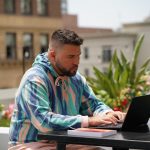 college student laptop study - Edify | Blogs Man in a pastel striped hoodie typing on a laptop at an outdoor terrace with a book on the table beside him, plants in the background.