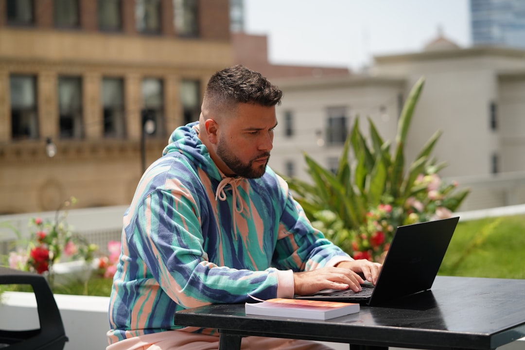 Man in a pastel striped hoodie typing on a laptop at an outdoor terrace with a book on the table beside him, plants in the background.