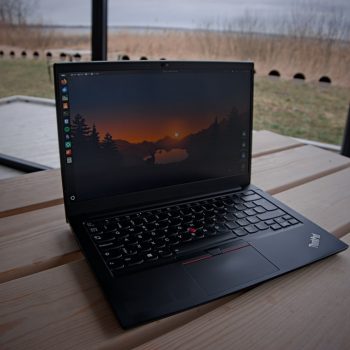 Lenovo ThinkPad laptop open on a light wooden table by a large window, with a lake and bare trees visible outside and sunset wallpaper on screen