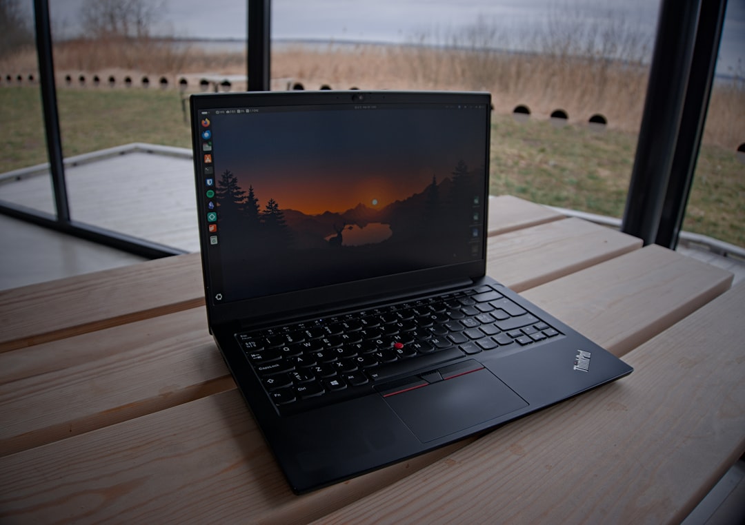 Lenovo ThinkPad laptop open on a light wooden table by a large window, with a lake and bare trees visible outside and sunset wallpaper on screen