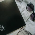 Overhead view of a closed black laptop with a white dragon logo, a white face mask, and black sunglasses with purple lenses on a light cloth surface.