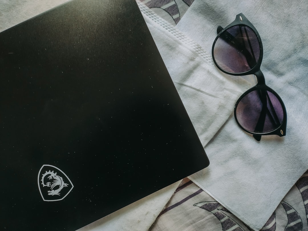 Overhead view of a closed black laptop with a white dragon logo, a white face mask, and black sunglasses with purple lenses on a light cloth surface.