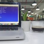 Open MacBook Pro on a white desk displaying code, with a small potted plant and a glass mug of black coffee nearby in a bright office space that’s out of focus.