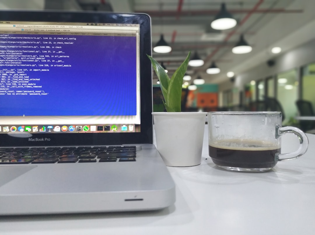 Open MacBook Pro on a white desk displaying code, with a small potted plant and a glass mug of black coffee nearby in a bright office space that’s out of focus.