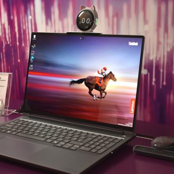 Lenovo ThinkBook laptop on a purple desk displaying a horse rider wallpaper, with a cat-ear webcam perched on top and accessories nearby.