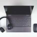 Top-down view of a gray laptop on a white desk, with black over-ear headphones resting on the keyboard and a wireless mouse to the right.