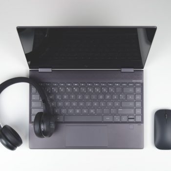 Top-down view of a gray laptop on a white desk, with black over-ear headphones resting on the keyboard and a wireless mouse to the right.