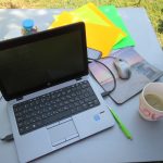Silver laptop on a gray table outdoors with a mouse, colorful paper sheets, a green pen, a coffee cup, and greenery in the background
