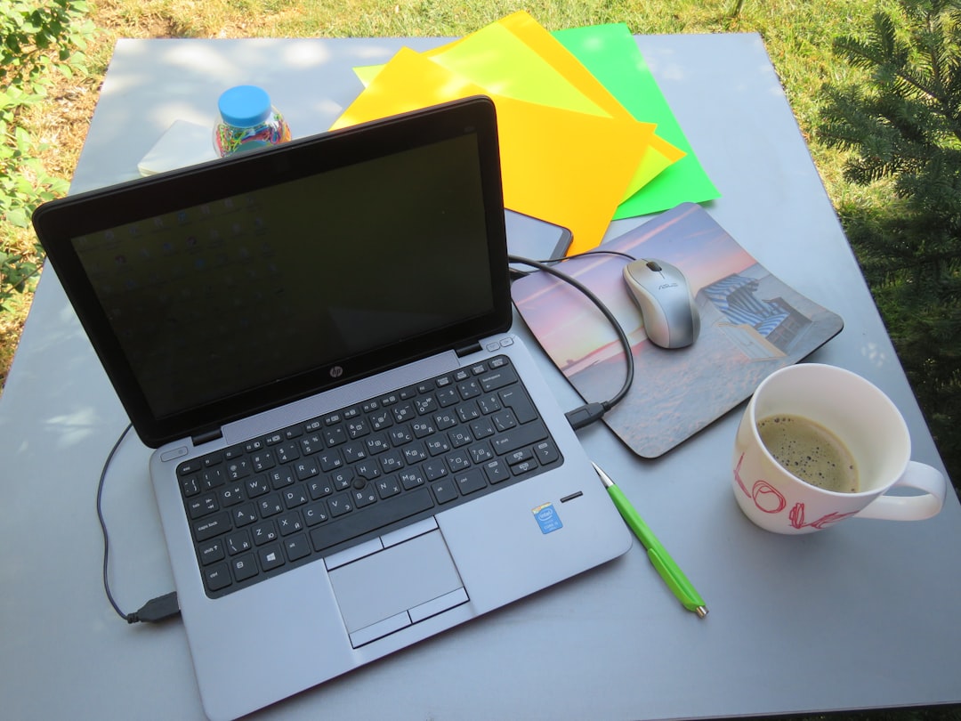 Silver laptop on a gray table outdoors with a mouse, colorful paper sheets, a green pen, a coffee cup, and greenery in the background
