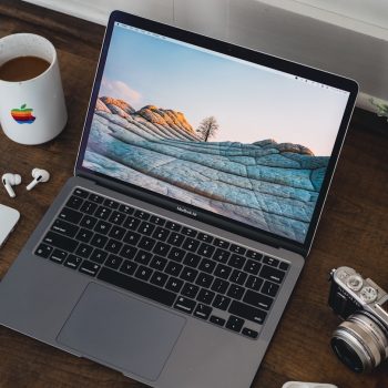 MacBook Air on a wooden desk with a rocky landscape wallpaper, surrounded by a coffee mug, iPhone, wireless earbuds, a vintage camera, and a potted plant.