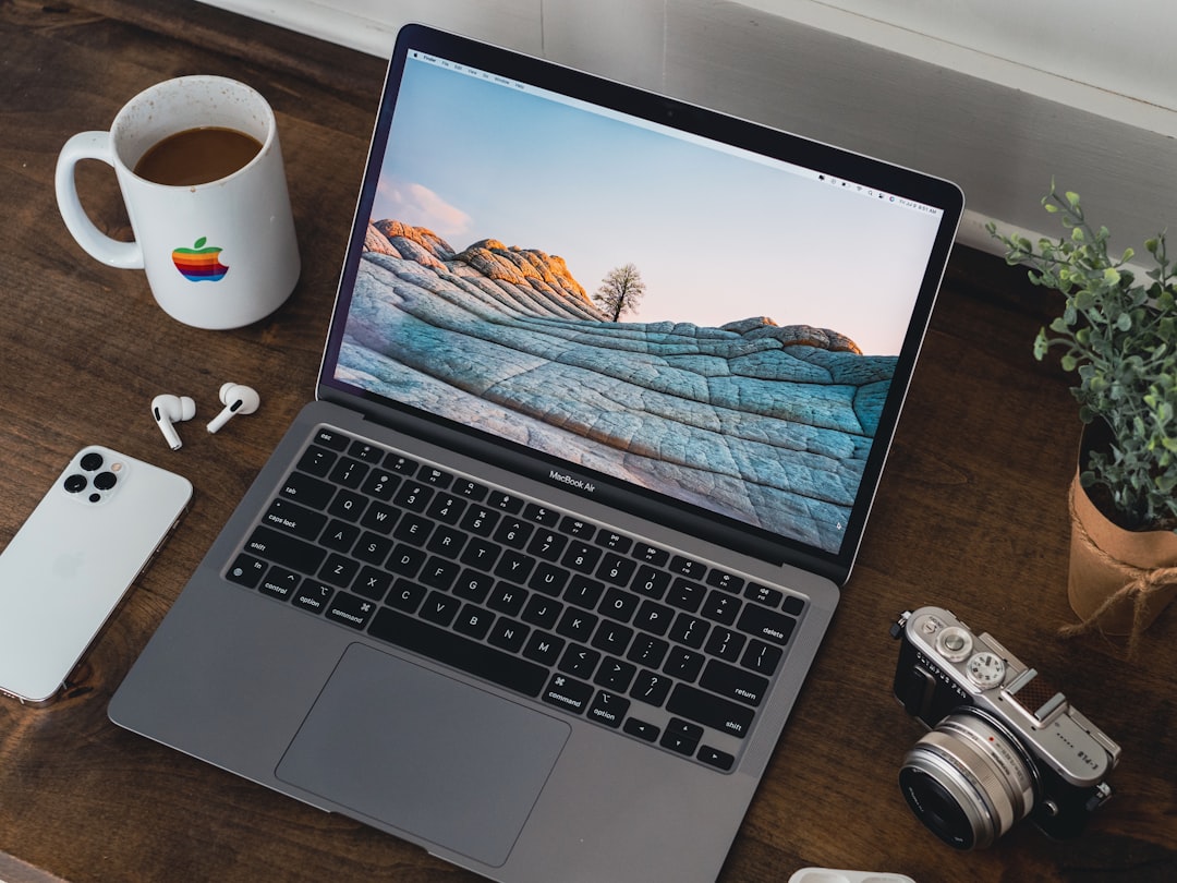 MacBook Air on a wooden desk with a rocky landscape wallpaper, surrounded by a coffee mug, iPhone, wireless earbuds, a vintage camera, and a potted plant.