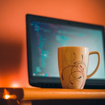 Ceramic mug with swirly design on a desk in front of a laptop displaying code, warm orange lighting.