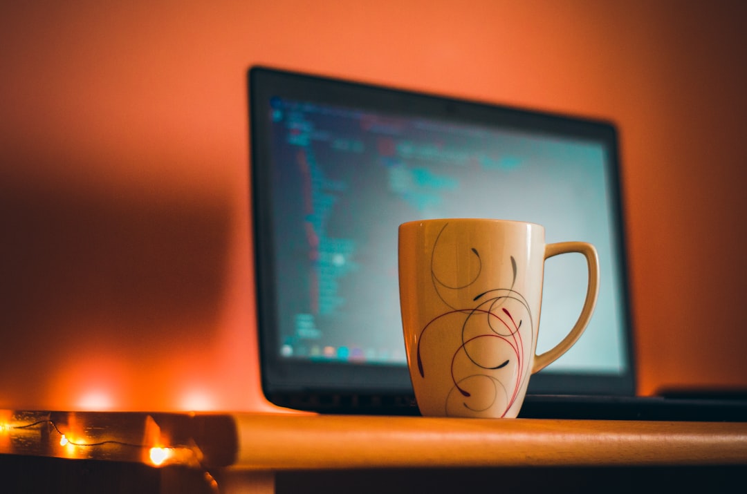 Ceramic mug with swirly design on a desk in front of a laptop displaying code, warm orange lighting.